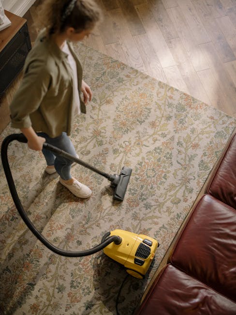 A person kneeling on a patterned rug in a domestic setting, using a yellow and black vacuum cleaner to clean a white cloth filter. The vacuum cleaner is positioned on a carpeted floor, with the lid op
